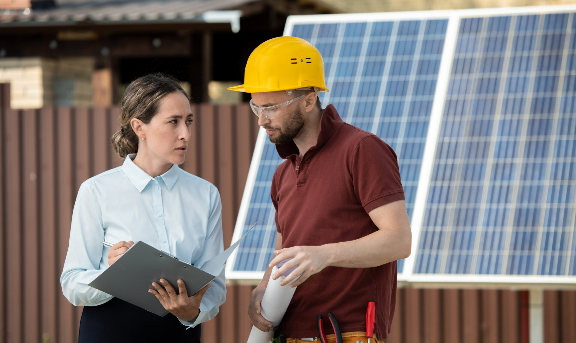 woman holding a clipboard talking to a man in a hard hat and safety glasses with solar panels behind them